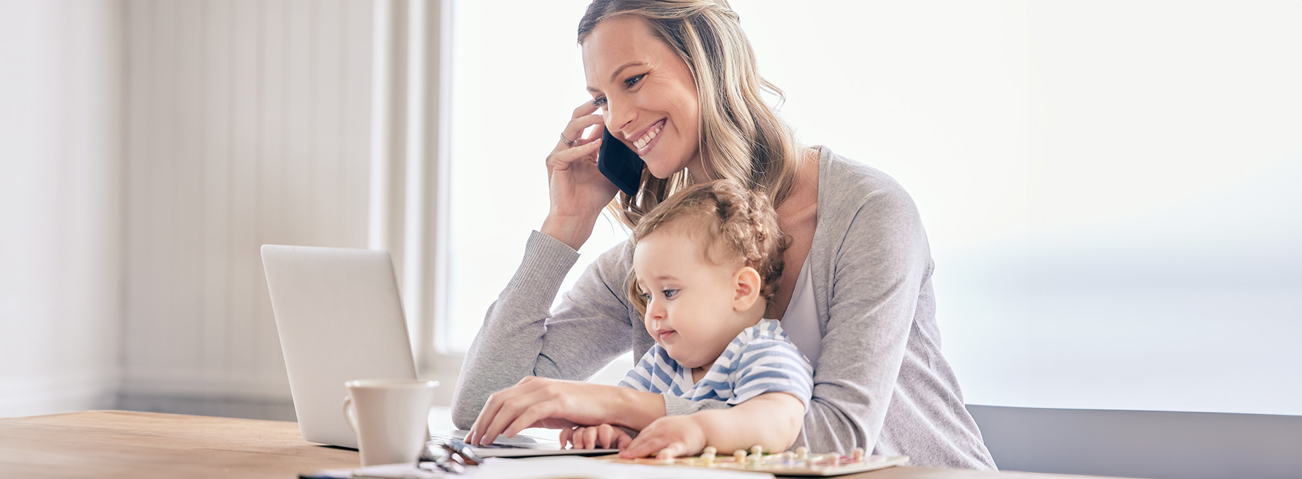 A woman and child are seated at a table with a laptop, engaged in an activity, while another person talks on the phone.
