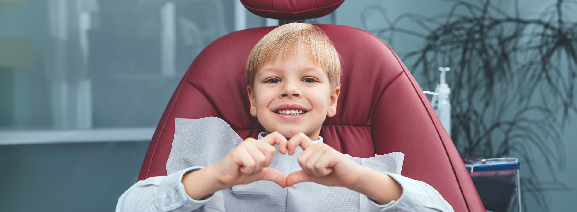 The image shows a young child sitting in a dental chair with their hands forming a heart shape above their head, smiling at the camera.