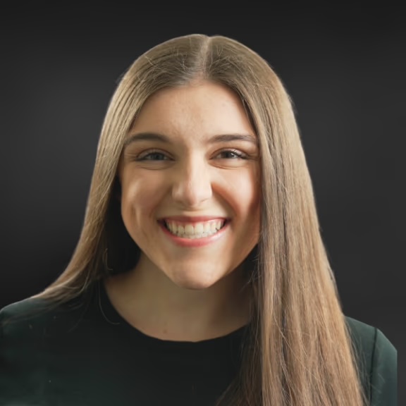 The image shows a person with long hair smiling at the camera, wearing a dark top against a blurred background.