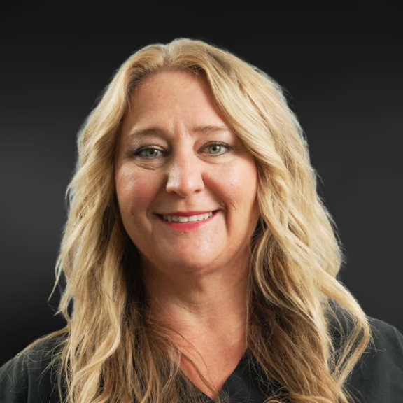 The image shows a woman with blonde hair, wearing a black top, posing for a portrait against a dark background.