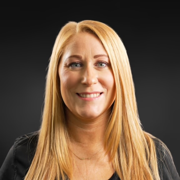 The image shows a woman with long blonde hair smiling at the camera. She has a light complexion and appears to be wearing makeup, including lipstick. Her attire includes a dark top with a visible necklace, and she has a serious expression on her face.