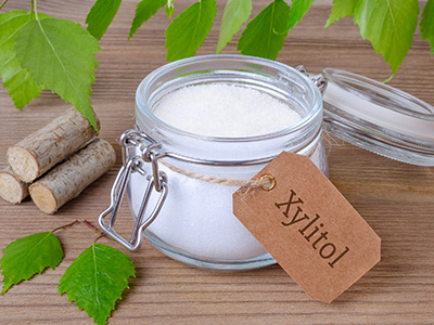 The image shows a clear glass jar with a label marked  xylitol,  containing a white powdery substance, placed on a wooden surface next to a bunch of green leaves, and accompanied by two cinnamon sticks tied together with string.