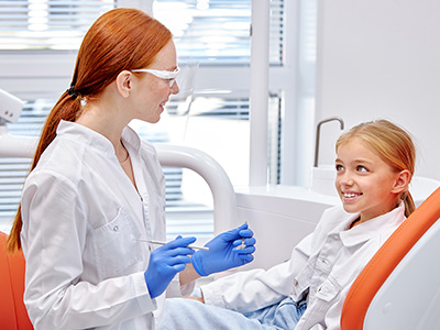 A dental professional attends to a young girl in a dental setting, with both individuals wearing blue gloves and face masks.
