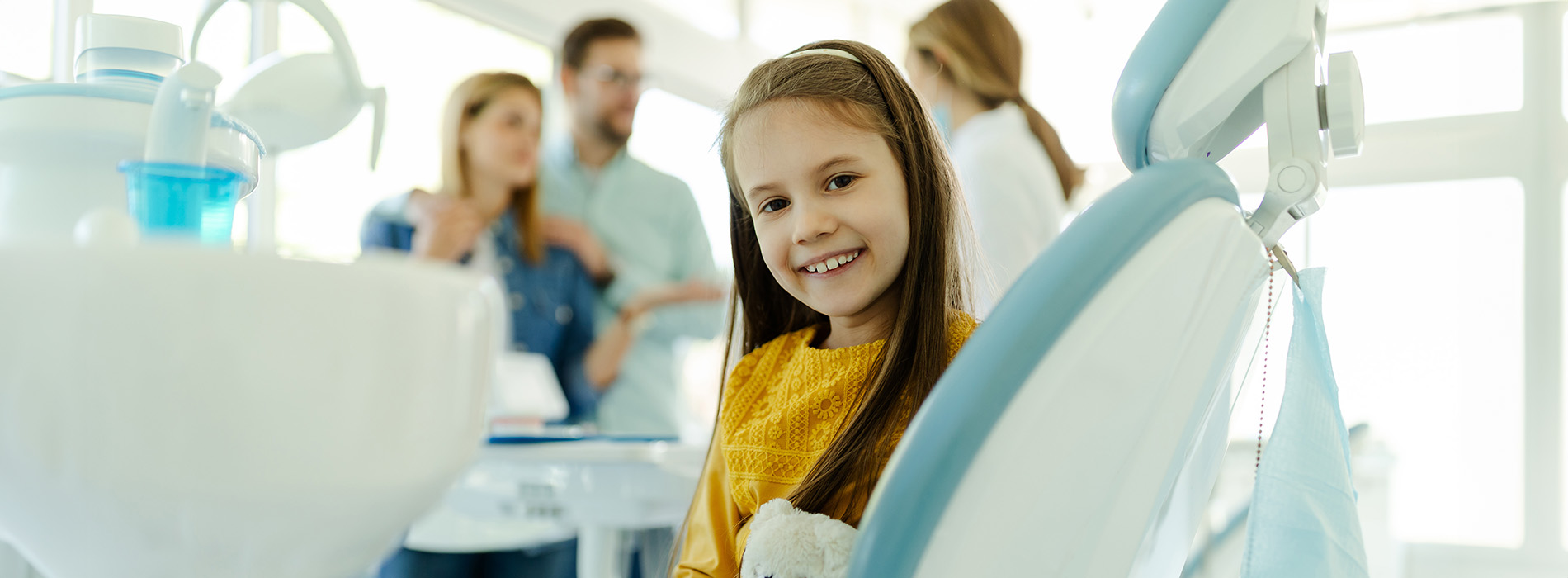 A young girl sitting in a dental chair with a backdrop of a dental office, surrounded by adults who appear to be dentists or staff.