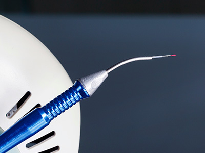 The image displays two close-up photographs of a blue dental tool with a needle-like tip, which appears to be a dental implant screw, being inserted into a white object that could be a model jaw or a simulated tooth socket.