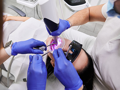 The image shows a dental professional performing a dental procedure on a patient, with two people wearing blue gloves and a bright light illuminating the area of work.