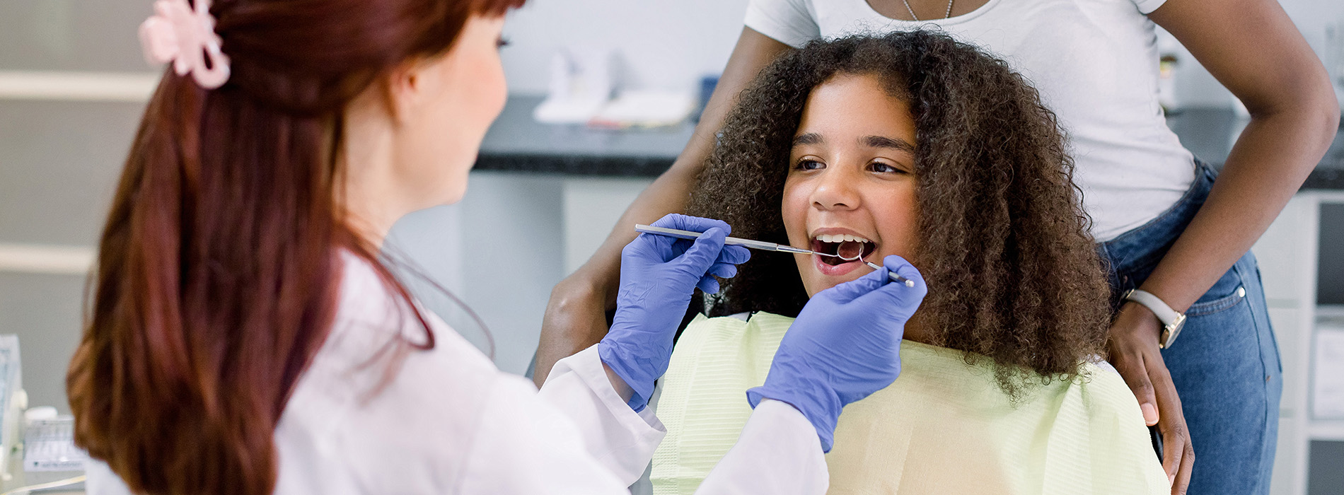 A dental hygienist is performing a cleaning procedure on a young girl s teeth with the assistance of a dentist, who is observing the process closely.