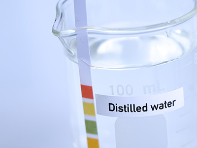 The image shows a clear glass test tube with a small amount of liquid inside, placed on a white surface, with a label indicating  distilled water  and measurement markings for volume in milliliters.