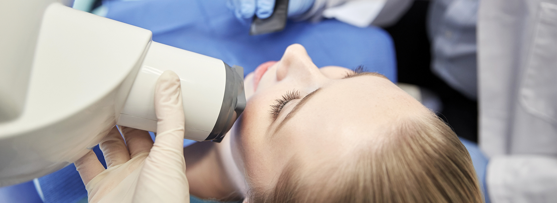 A person receiving medical treatment with a device, likely a microscope or similar instrument, held up close to their face by a healthcare professional, set against a blue background that suggests a clinical environment.