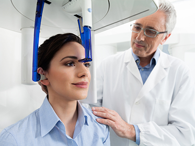In the image, there s a woman seated with her head inside a blue machine, possibly an MRI or similar diagnostic equipment, while a man in a white lab coat stands beside her, observing the process.